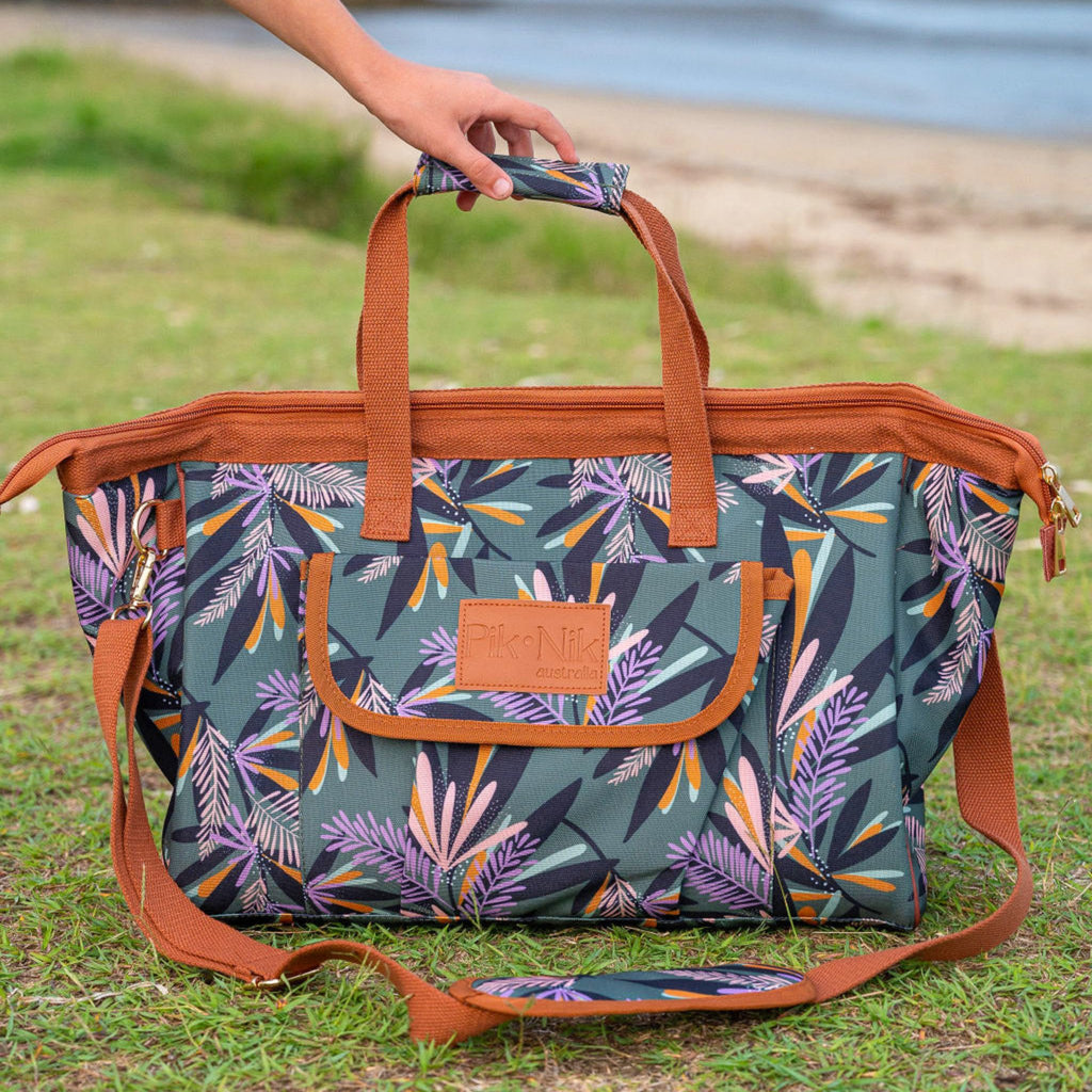A picnic cooler bag with a multicolored floral pattern set against a grassy background, featuring a leather logo and sturdy handles.